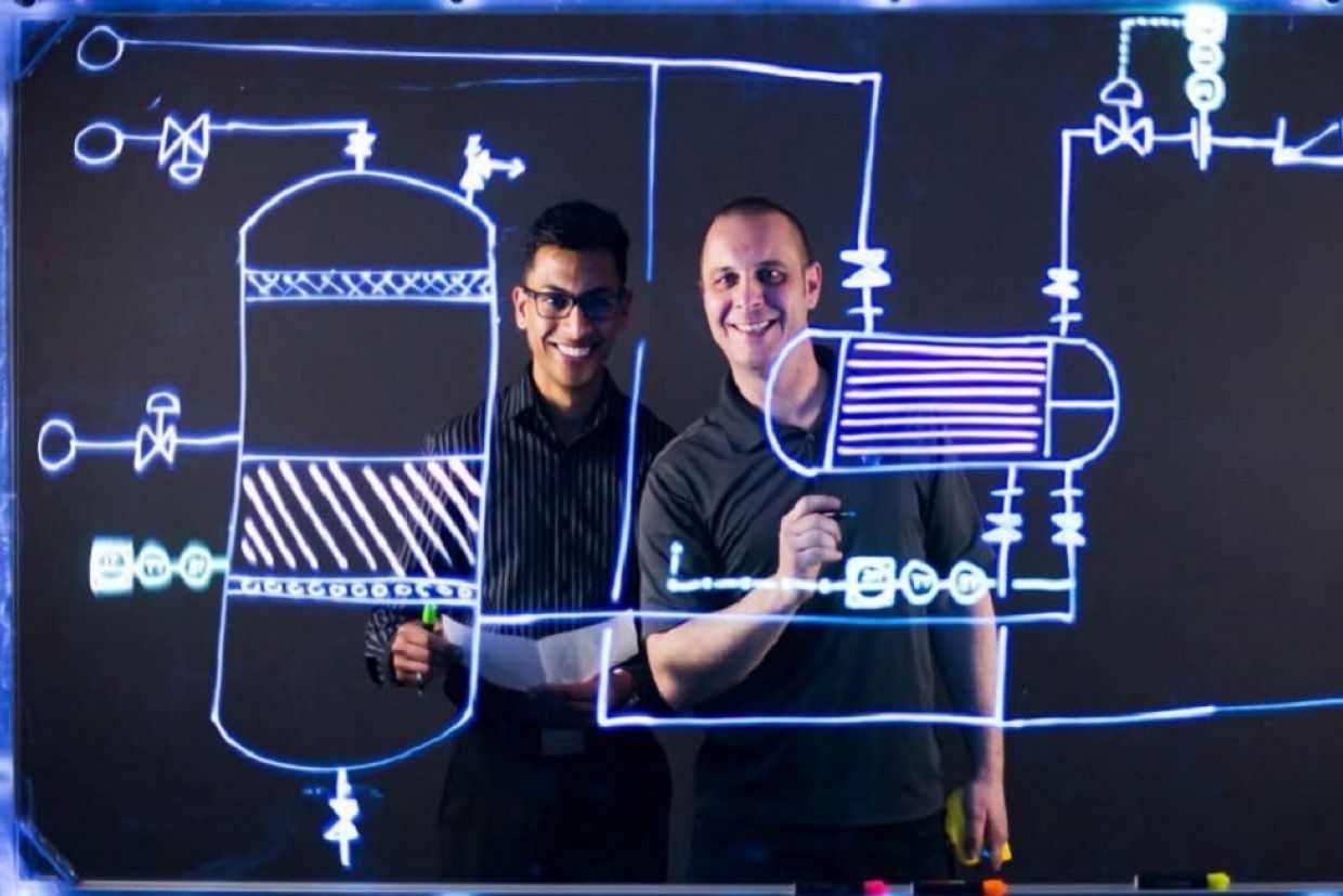 Two people smile from behind a clear whiteboard with schematics on it