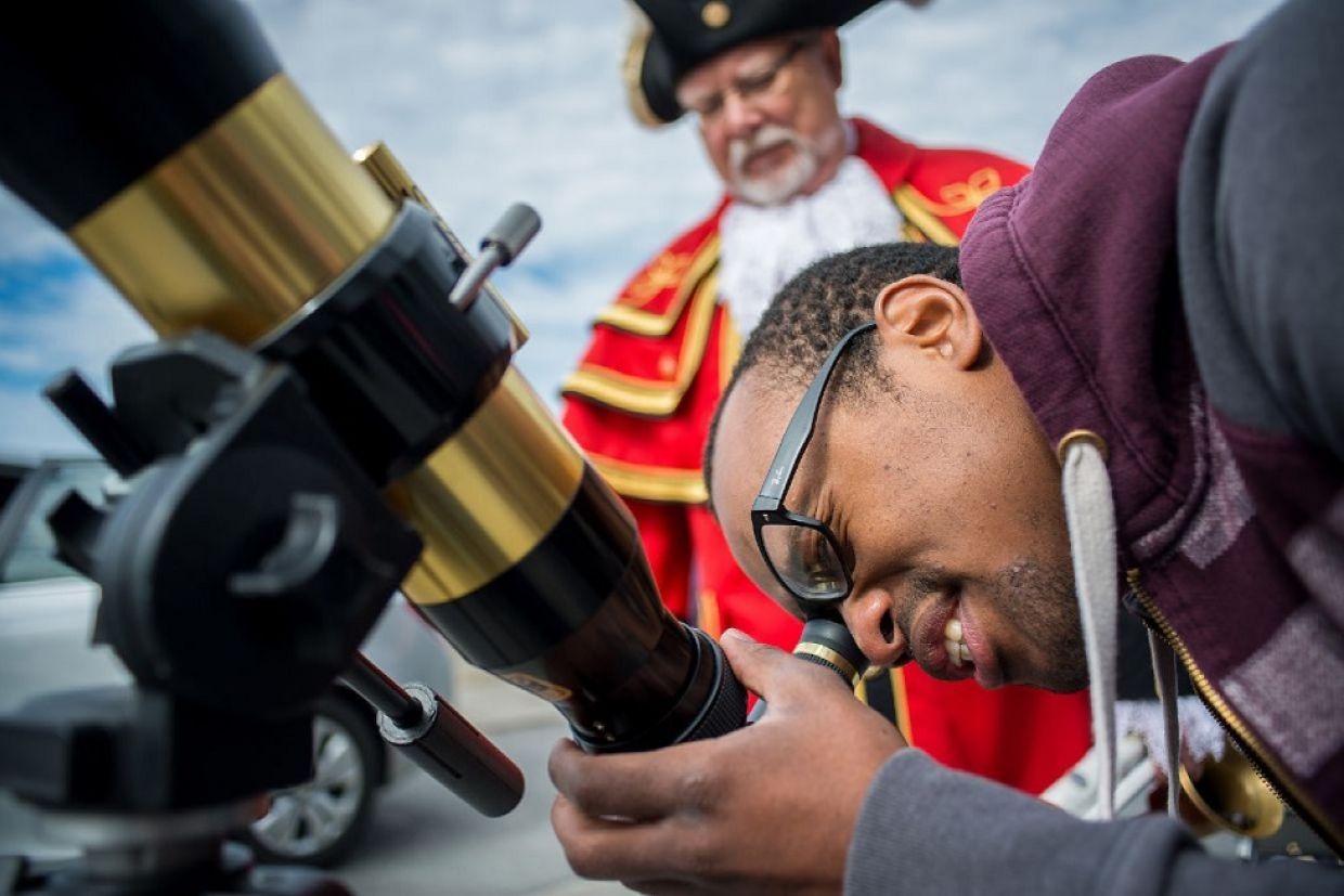 A man with a concentrated expression looks through telescope as Kingston town crier looks on