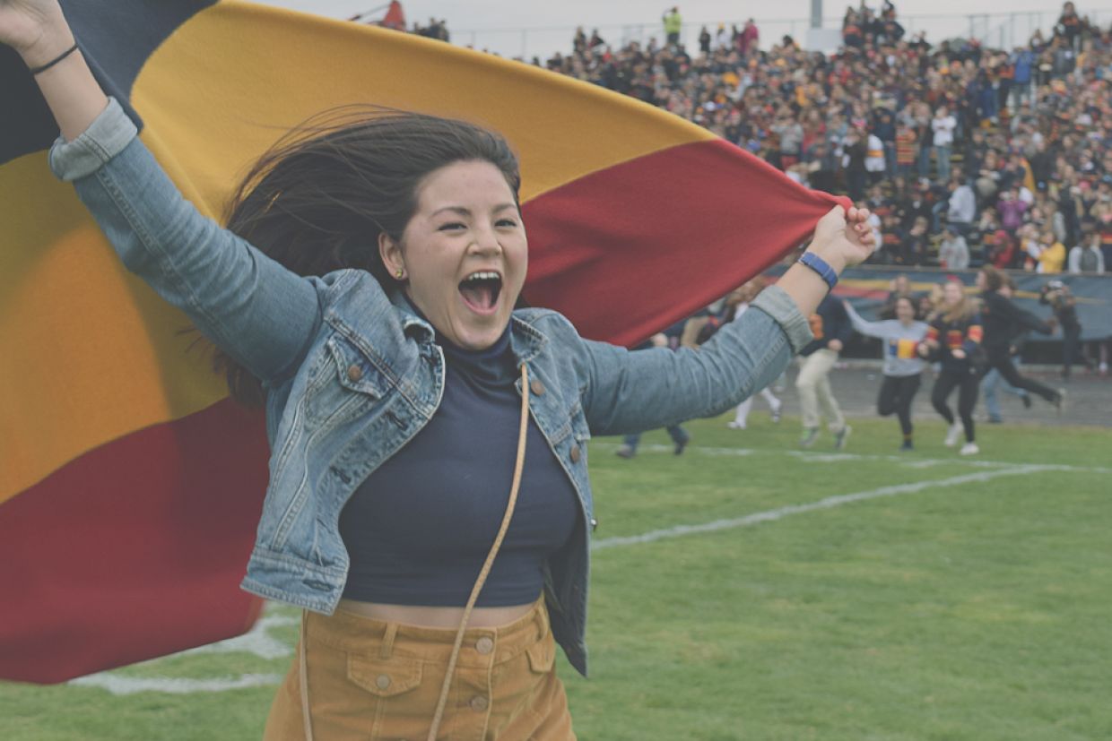 Woman running across football field holding an alumni flag over her head.
