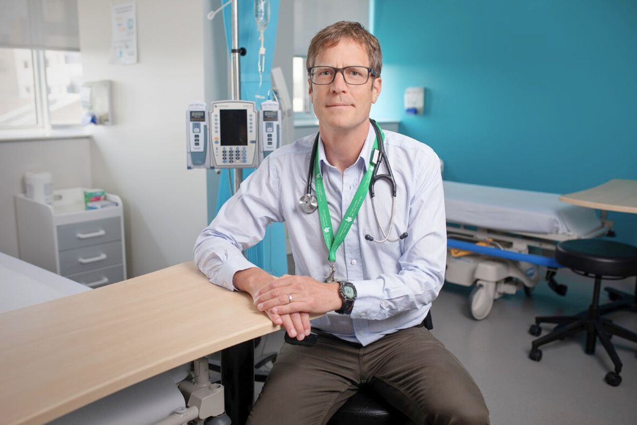 A doctor with a stethoscope and ID badge sits on a hospital bed's edge. The room has medical equipment, a bright blue wall, and an open, professional vibe.