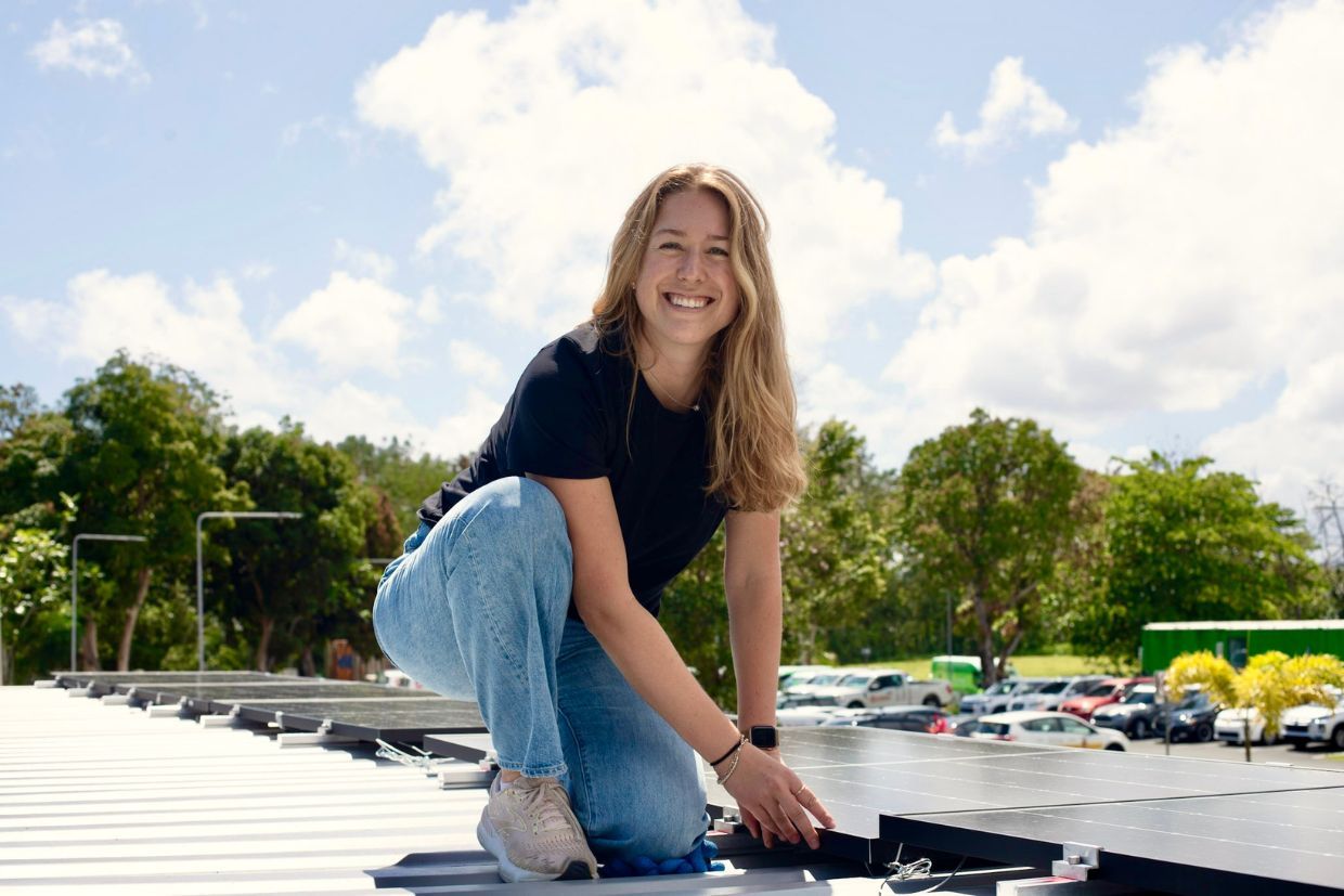 Meghan Wood, Com’18, smiles while kneeling on a rooftop showcasing an all-in-one solar battery pod.