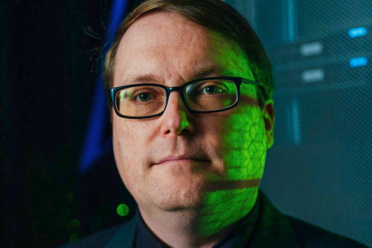 Ryan Grant, wearing glasses and a suit, stands before a computer, highlighting his contributions to supercomputing advancements.