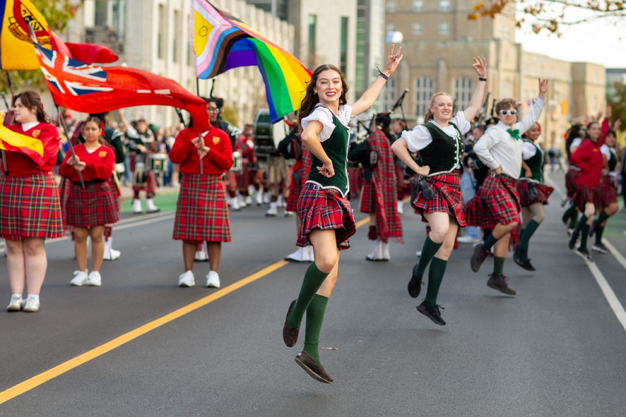 A group of Queen's students in colorful kilts joyfully dancing together on a lively street during a Homecoming celebration.