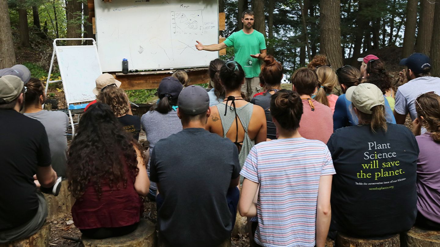 A man presents to an audience, gesturing towards a screen with visual aids, while attendees listen attentively.