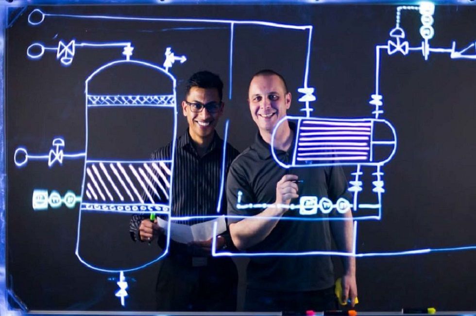 Two people smile from behind a clear whiteboard with schematics on it