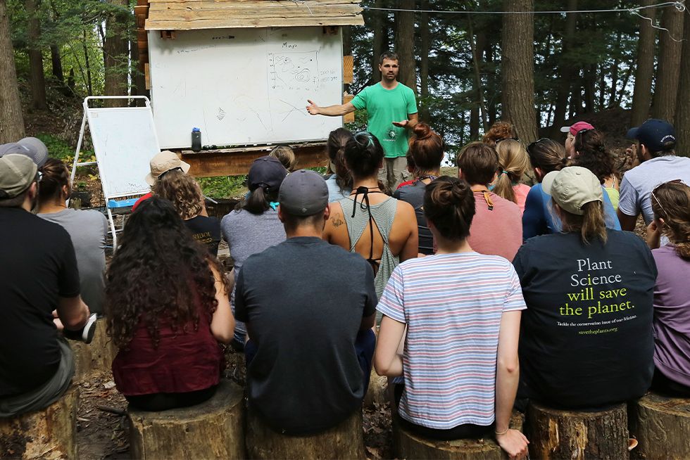 A man presents to an audience, gesturing towards a screen with visual aids, while attendees listen attentively.