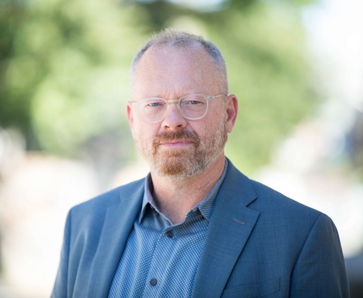 Headshot of Christopher Donaldson. He wears a blue suit and there is green foliage behind him. 