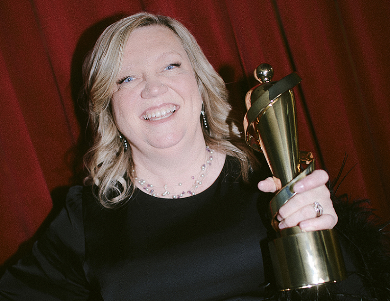 A smiling Jeannie Hunter holding her Juno, with a red curtain behind her. 
