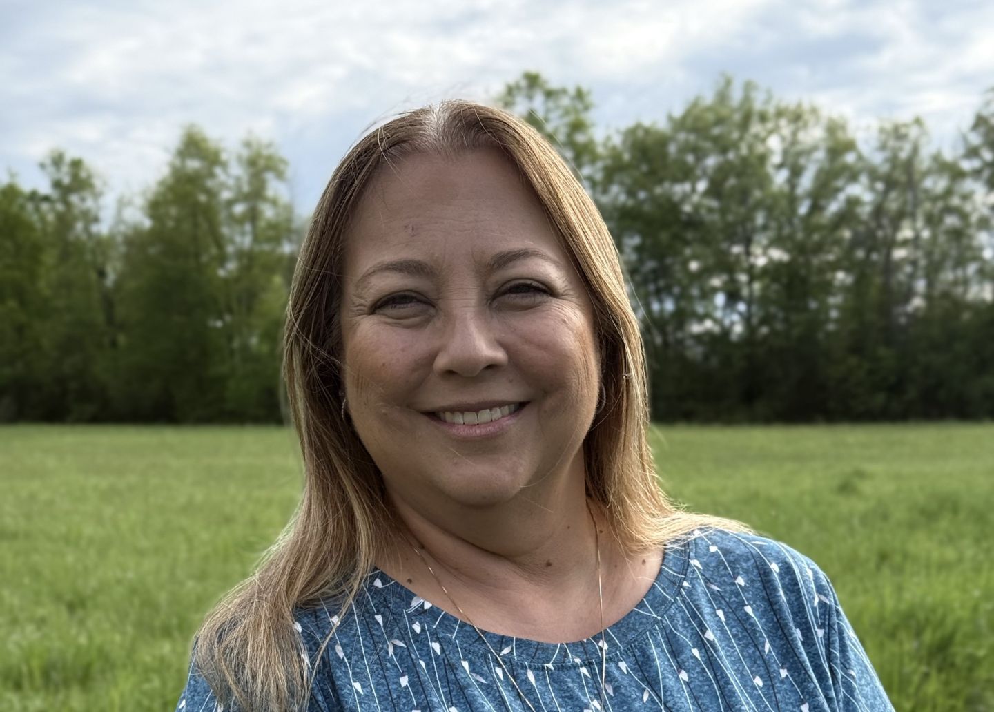 Lisa Maracle smiling, wearing a blue shirt, with a green-grass field behind her and trees beyond that.