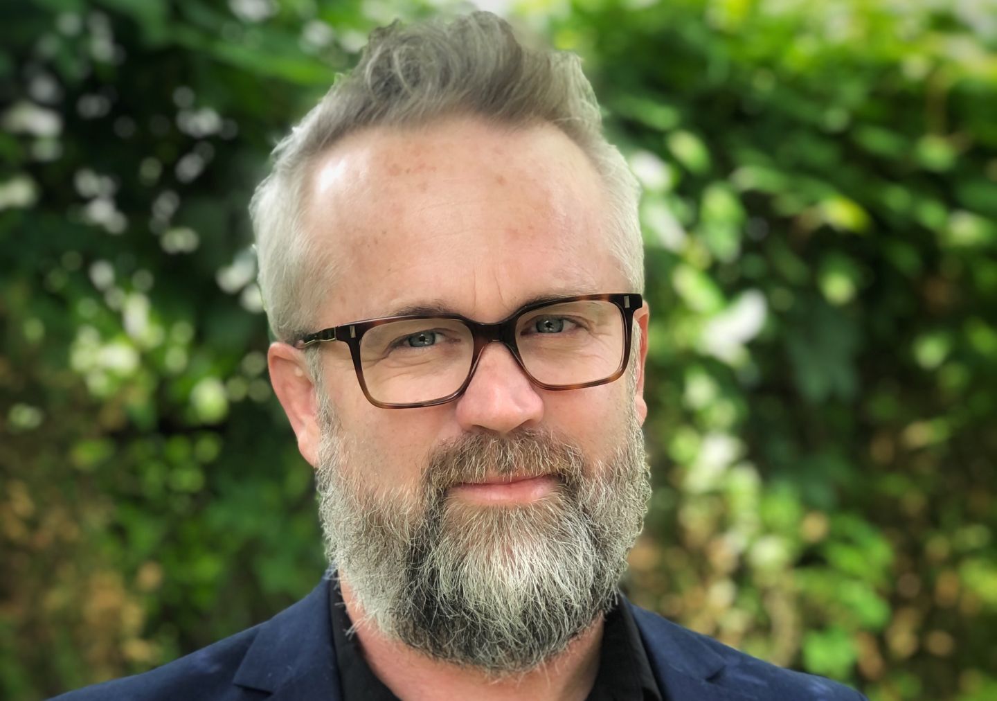Headshot of Ron Shore wearing a blue jacket and dark glasses, with green vegetation behind him.