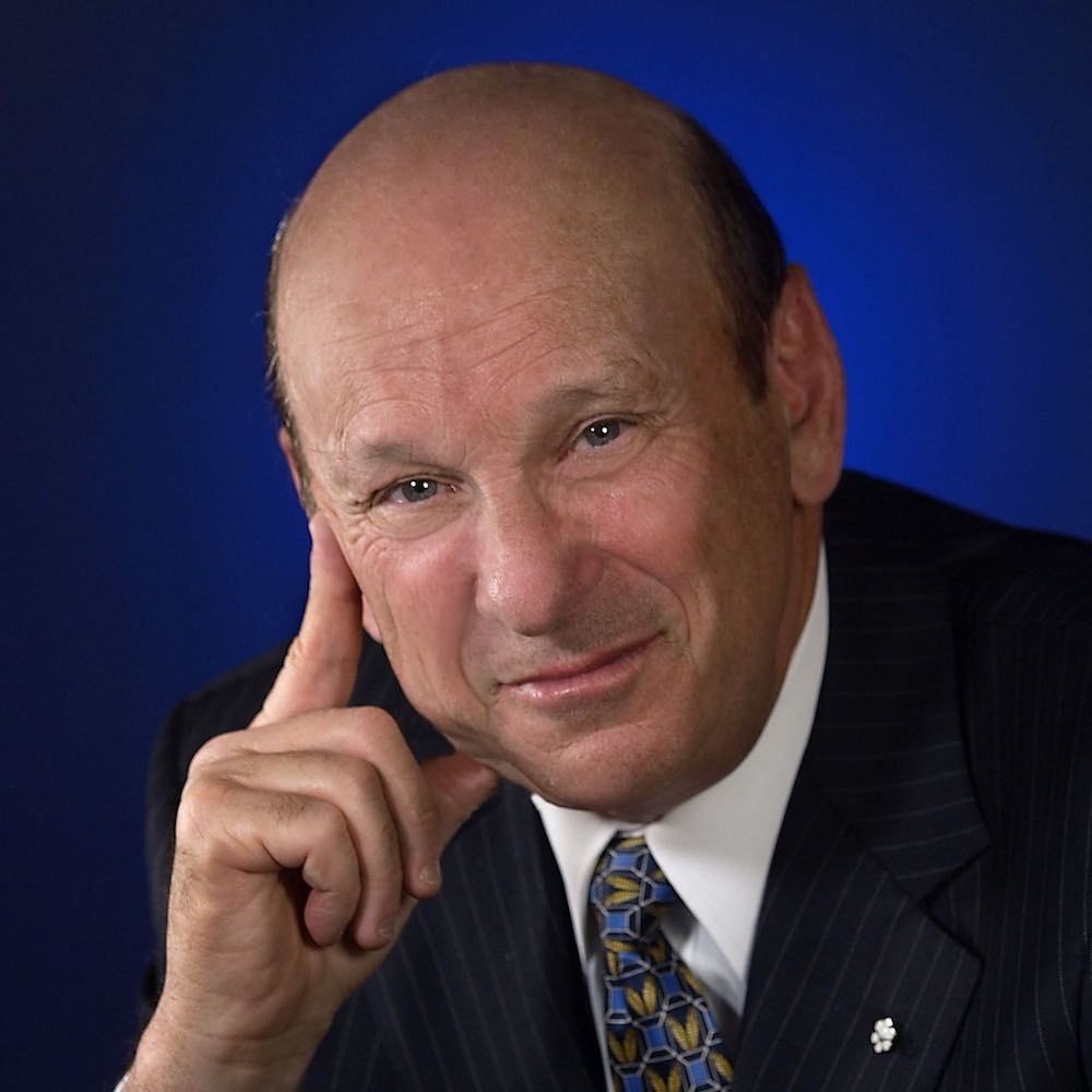 Headshot of Seymour Schulich smiling, wearing a suit and a tie, with a dark blue background.