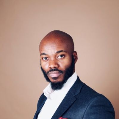 Akin standing with one arm folded infront of their stomach, Wearing a blue blazer with a white shirt and red pocket square