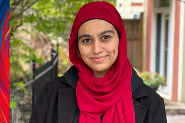 photo of Aliza Tiwana looking at the camera smiling, Aliza is wearing a red hijab and black jacket. Trees and building windows in the background
