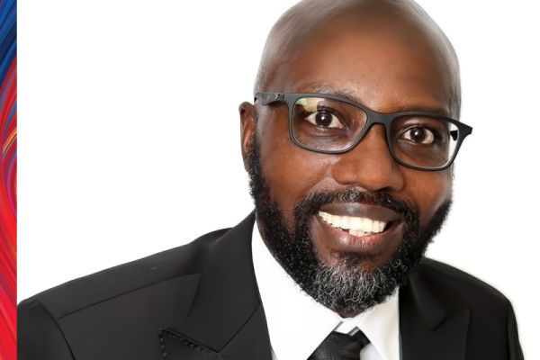 Norman Musengimana smiling wearing a black suit jacket, white shirt, black tie and black framed glasses