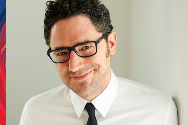 Elan Mastai smiling wearing a white collared shirt, black tie and black framed glasses