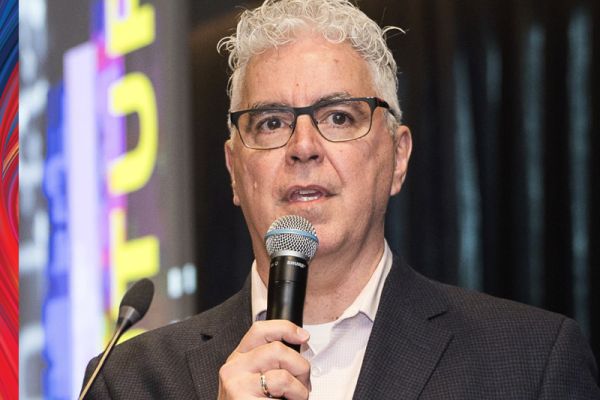 photo of David Phipps holding a microphone wearing a black suit jacket, collared shirt and black framed glasses