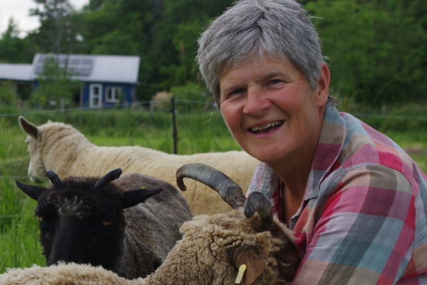 Lisa Webb stands with her goats on her farm, surrounded by green pastures and a clear sky.