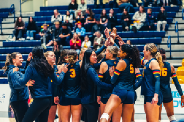 Queen's Women's Volleyball team celebrate victory in circle with arms in the air together