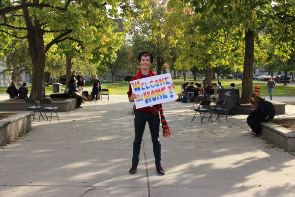 Student standing outside of Grant Hall smiling, holding a Tricolour sign that reads "Welcome Home"