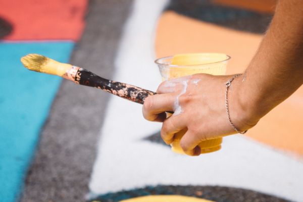 An artist holds a paintbrush, and cup of paint over a mural on the ground.