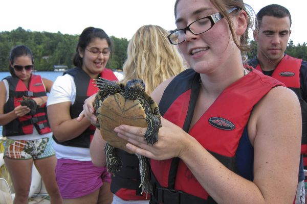 A young person in a life vest holds a painted turtle and smiles. I group of students in the background are holding turtles and smiling.