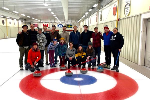 A group of alumni, family, and friends posed in front of the button on a curling rink