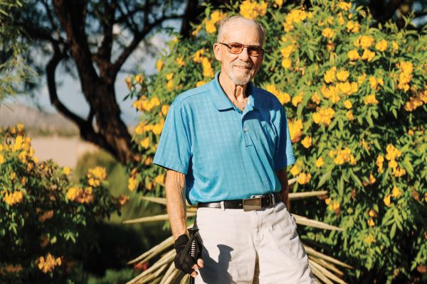 John Simpson is wearing a blue shirt and khaki pants, standing in front of a bush with yellow flowers.