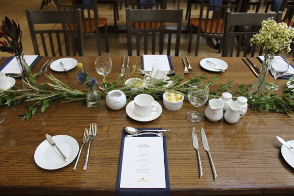A formal place setting on wooden table with a flower centre piece.