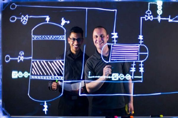Two people smile from behind a clear whiteboard with schematics on it