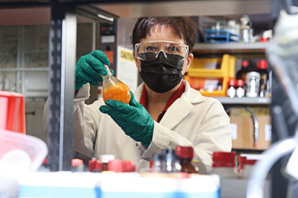 Cathleen Crudden in a lab coat, goggles, and mask holds a science beaker. 