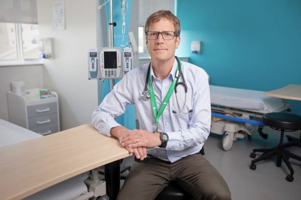 A doctor with a stethoscope and ID badge sits on a hospital bed's edge. The room has medical equipment, a bright blue wall, and an open, professional vibe.