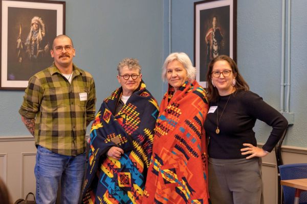 Group of four individuals posing happy to the camera with a grey background. Two of them, wearing beautiful indigenous pieces of clothing.