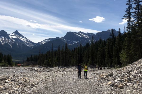 Two people walk on rocky ground with trees and mountains in the background
