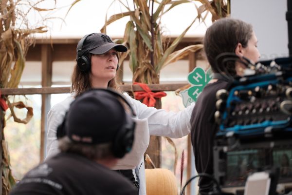 Katie Uhlmann on the set of Cows Come Home, wearing a dark hat and headphones, with corn husks behind her. 