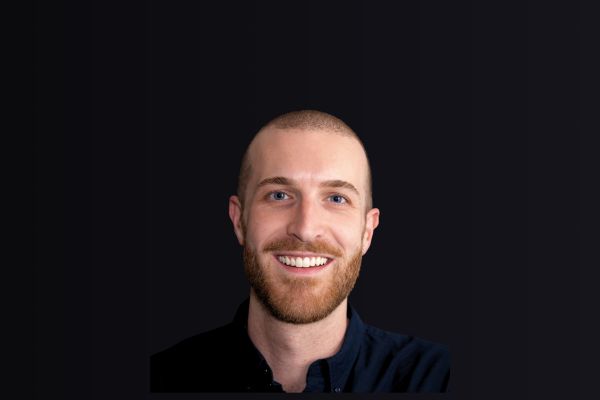 A smiling Sean McCabe wearing a dark-collared shirt. The background is black. 
