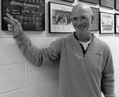 Black and white photo of Stuart Archibald standing in a school hallway and pointing at a photo on the wall.