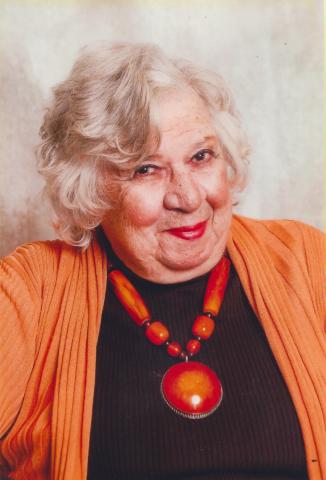 Portrait of older woman with blonde hair wearing a sweater and cardigan, with a large beaded necklace.