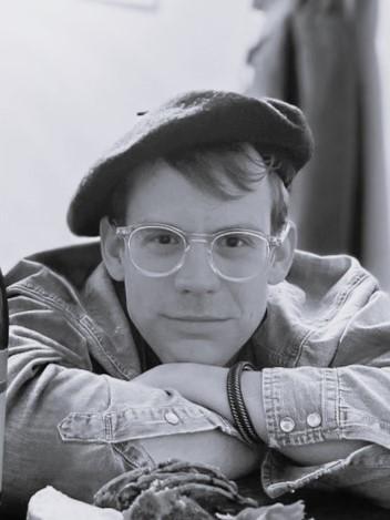 Photo of a man wearing a beret, glasses, and a jean shirt. His arms are folded in front of him with his chin resting on a hand.