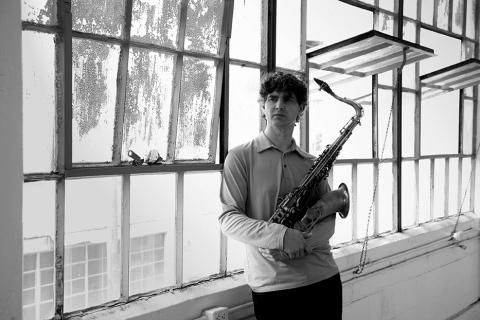 Andrew Rathbun leans against windows of an old warehouse, holding a saxophone to his chest.