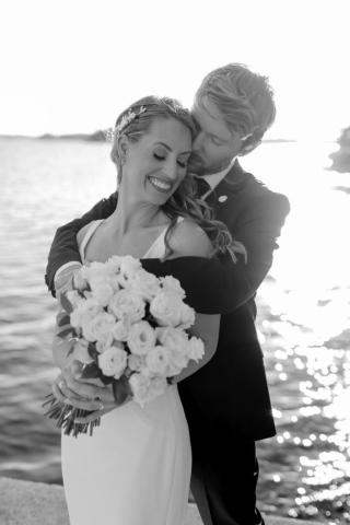 Ryan O'Callaghan hugs his wife, Shea Wood, from behind. Shea is wearing a wedding dress and holding a bouquet of roses.