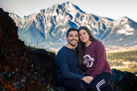 Ashley Drobot and fiancé sit on some rocks with a mountain in the background. Their bodies are turned towards each other with one hand on top of the others.