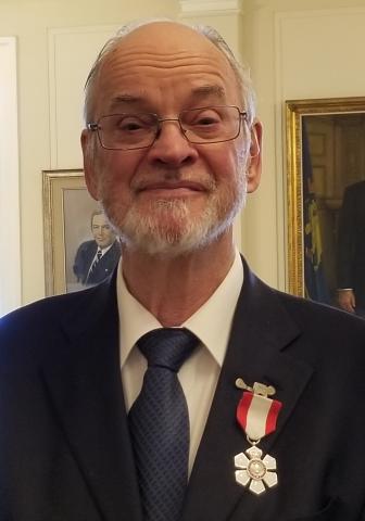 A man, with grey hair and a beard, wearing glasses and a suit, smiles at the camera. He has a medal pinned to his lapel and there are painted portraits in the background.
