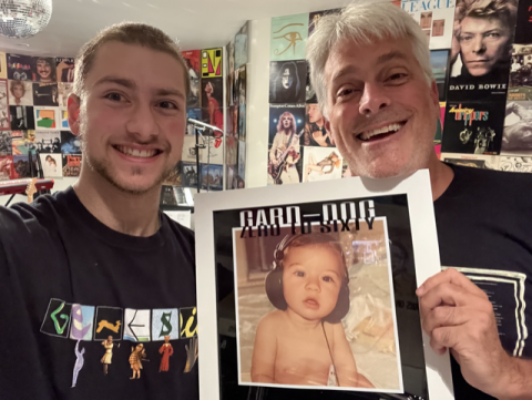 Colin Gardiner holds up a copy of his album cover which depicts a baby wearing headphones. Max Gardiner stands beside him with a wall covered in album covers behind them.