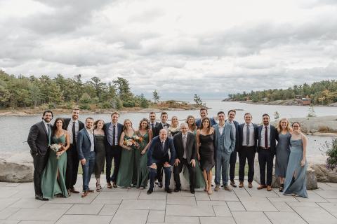 A large wedding party poses in front of a lake.