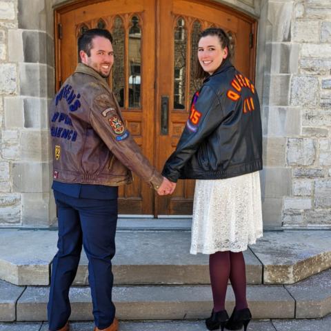 Jenna Dibblee and Connor Peebles stand in front of wooden doors on Queen's campus, wearing their faculty leather jackets. Their bodies face the door but their heads are turned around facing the camera. They are holding hands.