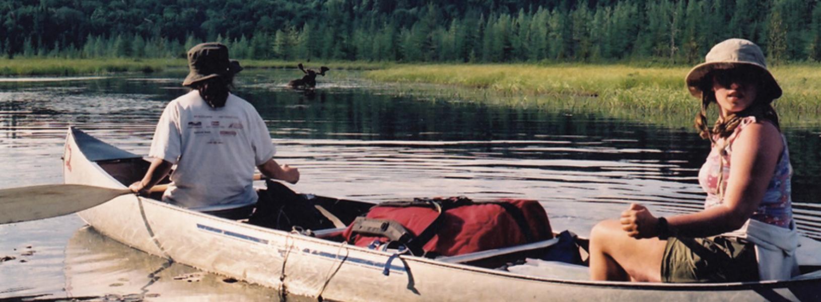 Two students paddle a canoe on a lake lined with evergreen trees.