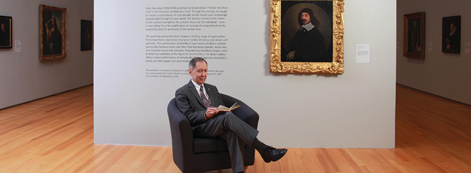Carlos Prado reads a book while sitting in a chair. Descartes as painted by Nason hangs on a wall behind him.
