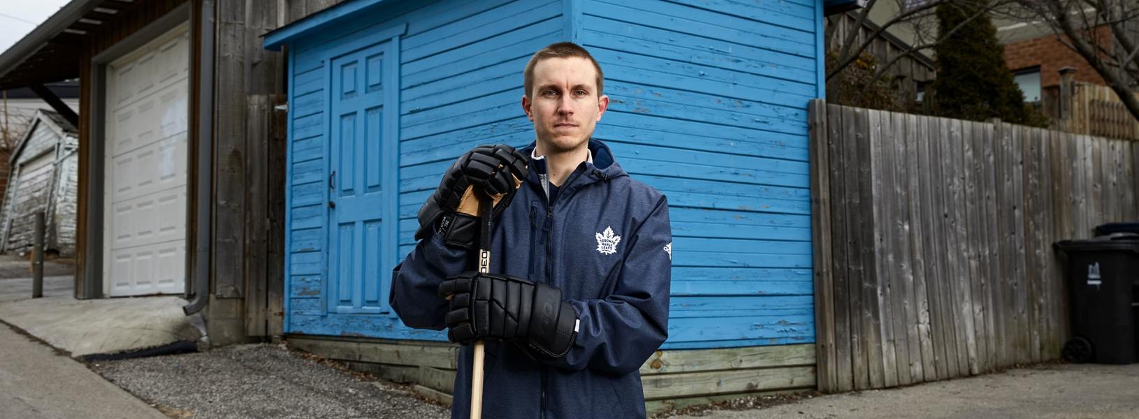 Darryl Metcalfe standing in front of a blue wooden shed holding a hockey stick and wearing hockey gloves.