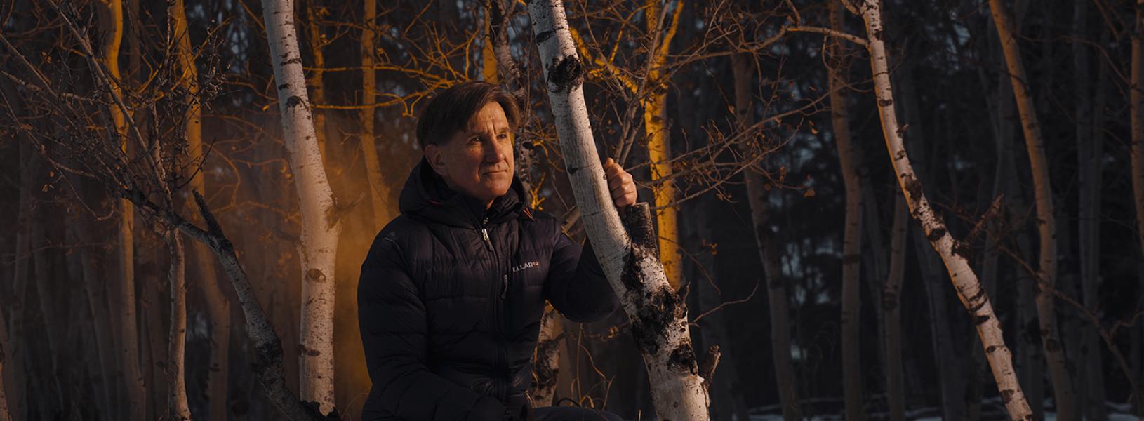 Edward Struzik is crouching in the woods wearing a winter jacket. He is surrounded by trees with a reflection of the light from a fire appearing behind him.