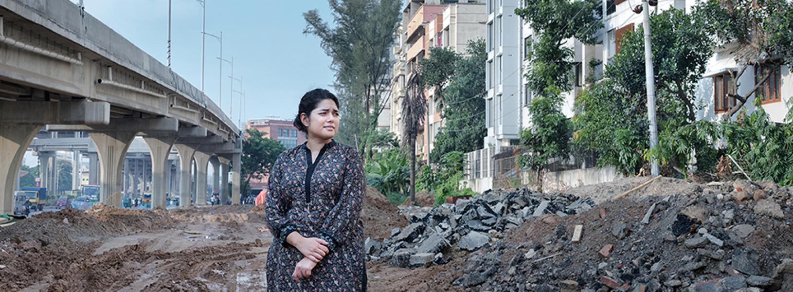 Bushra Afreen stands in mud and stones, rocks, and broken concrete beside an overpass and apartment buildings.